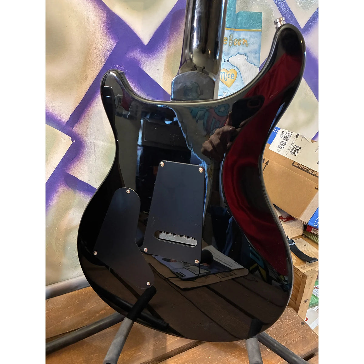 A glossy black electric guitar body, likely a Fender Strat-style model, photographed from the back on a stand in a cluttered indoor setting with cardboard boxes and a purple spray-painted background visible.