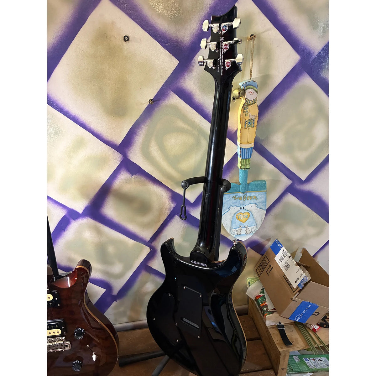 A black electric guitar with a glossy finish stands vertically against a patterned wall. The headstock is visible at the top, and another guitar is partially visible on the left, with some boxes on the floor in the background.
