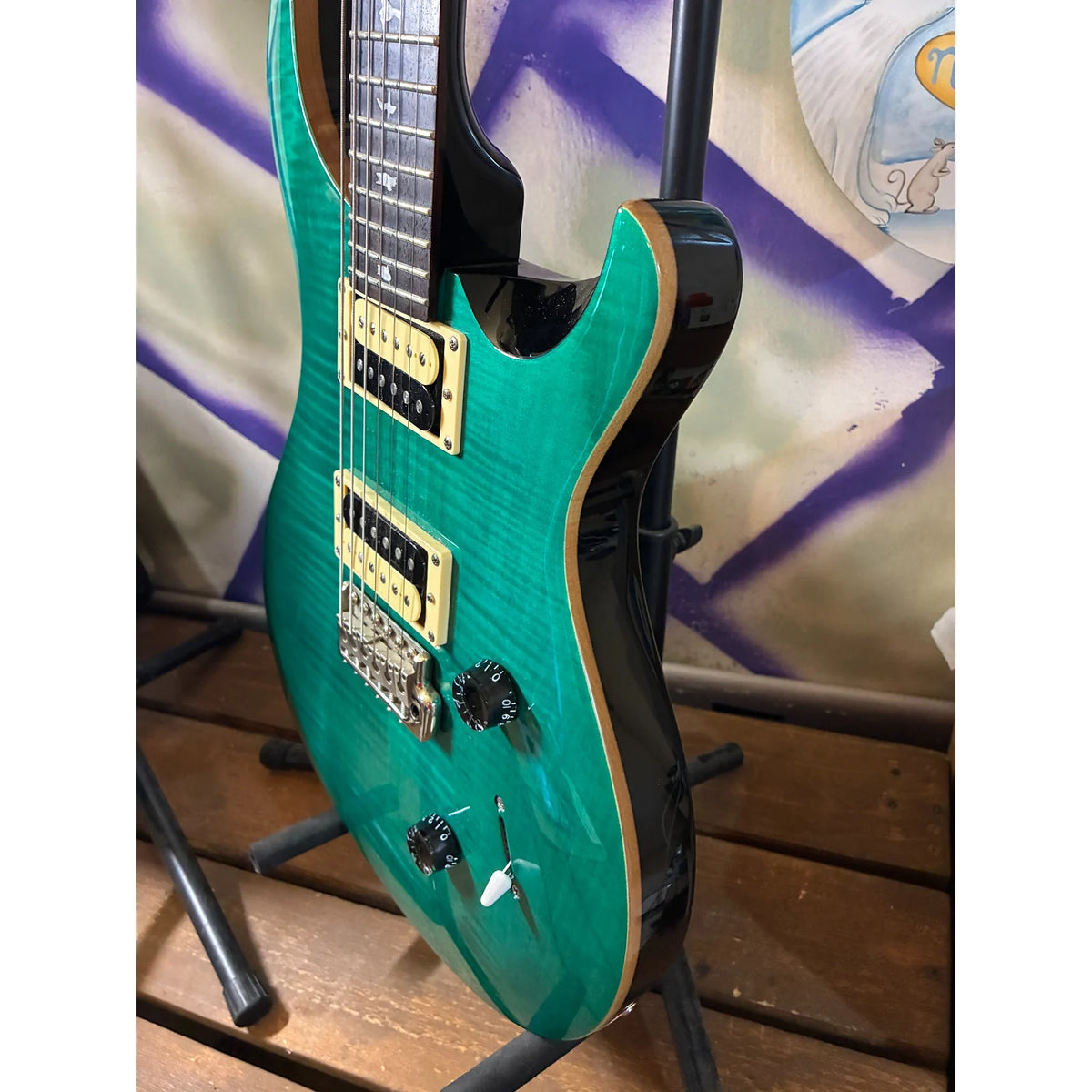 A turquoise green electric guitar with gold hardware and a black body, displayed on a stand in front of a purple and white patterned backdrop.