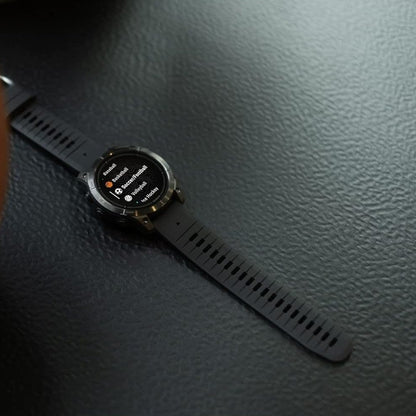 A black smartwatch with a round face and matching black strap, resting on a dark textured surface next to a brown leather object.