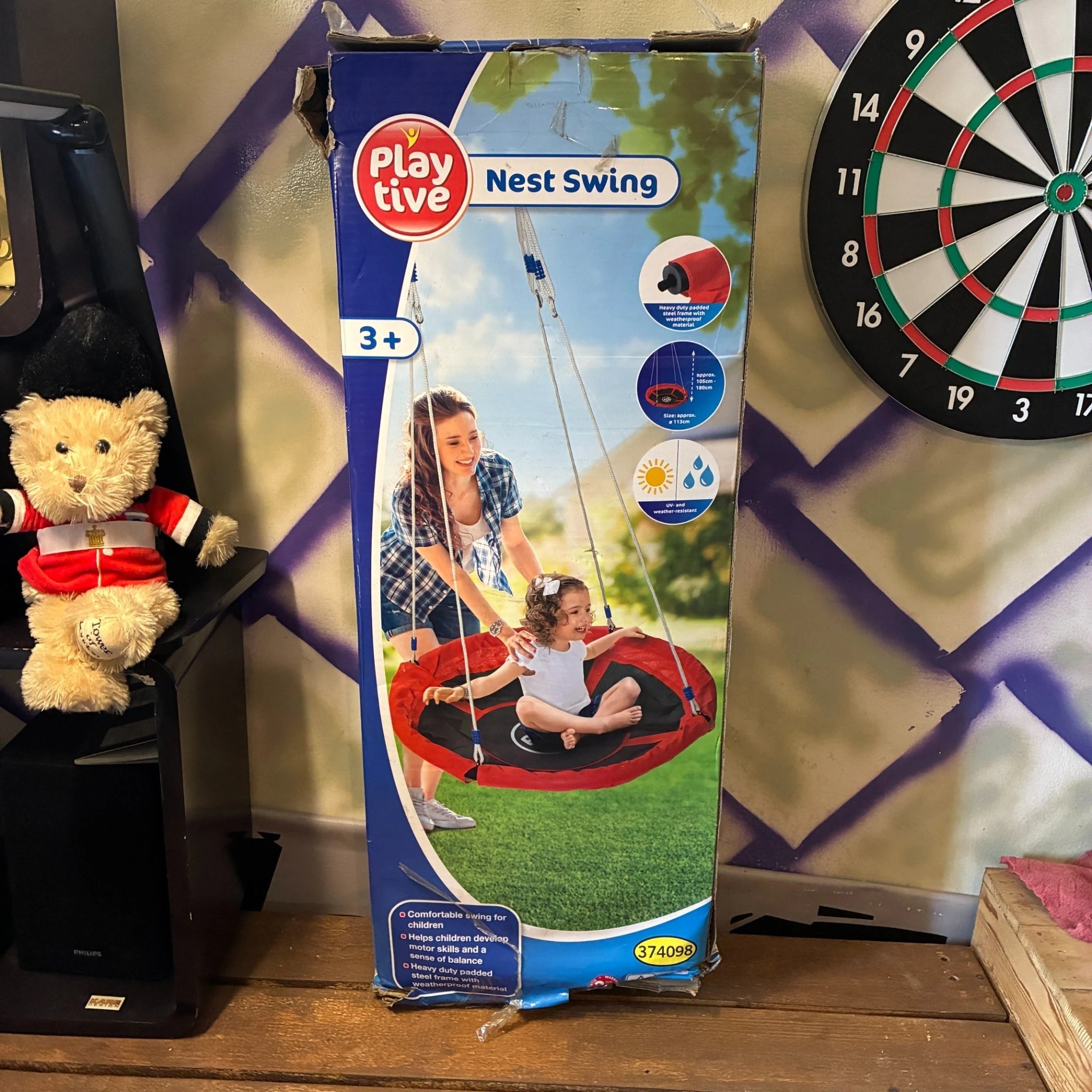 The retail packaging box for the Playtive Nest Swing, standing on a wooden shelf in a room. The box features a blue and white design with a photo of a woman and child using the red and black circular swing.