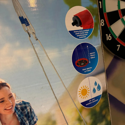Close-up of a product package or display panel showing three circular feature callouts: a heavy-duty padded slide frame with weatherproof material, a star rating graphic, and a sun-and-water weather-resistant icon. A partial person in a plaid shirt and a dartboard are also visible in the frame.
