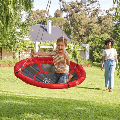 Child on a red and gray swing set in a backyard with a woman walking by.