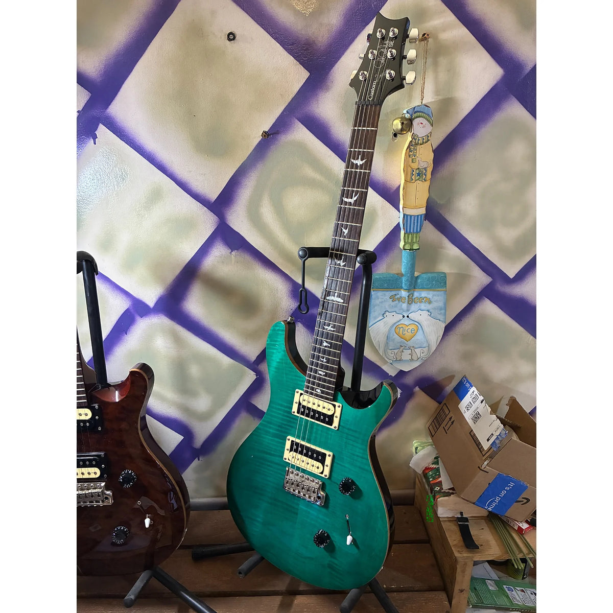 A teal electric guitar on a stand in a cluttered room with a purple and beige patterned wall, next to another guitar.