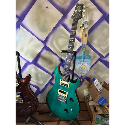A teal electric guitar on a stand in a cluttered room with a purple and beige patterned wall, next to another guitar.