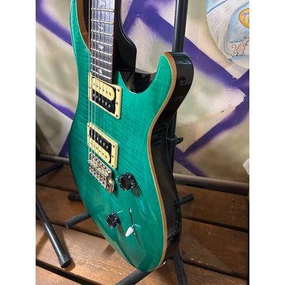 A turquoise green electric guitar with gold hardware and a black body, displayed on a stand in front of a purple and white patterned backdrop.
