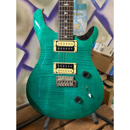 A turquoise green electric guitar with gold hardware and a dark fretboard, displayed on a stand in a cluttered room with cardboard boxes and a graffiti-patterned wall in the background.